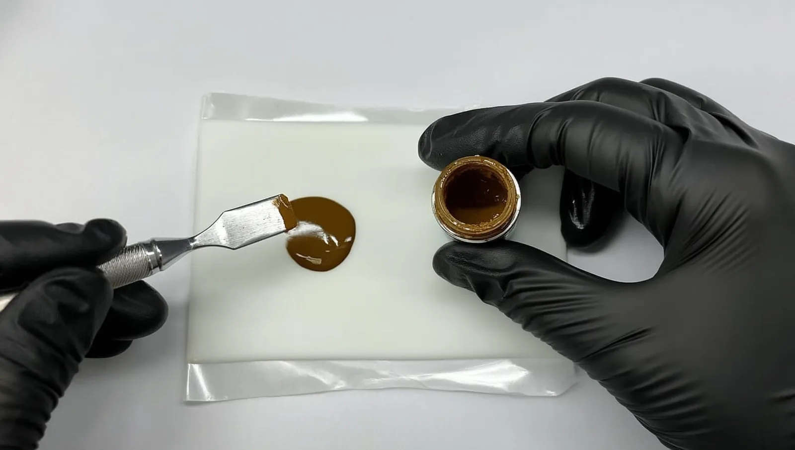 A microblading artist's gloved hand holding a small cap of brown iron oxide pigment beside a hand tool on a clean tray.