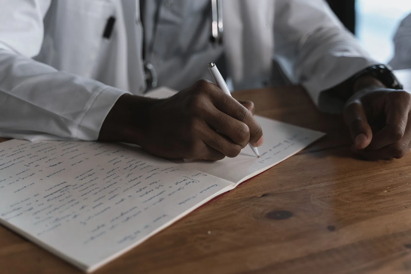 A doctor writing notes with a pen in an open medical notebook on a wooden desk.
