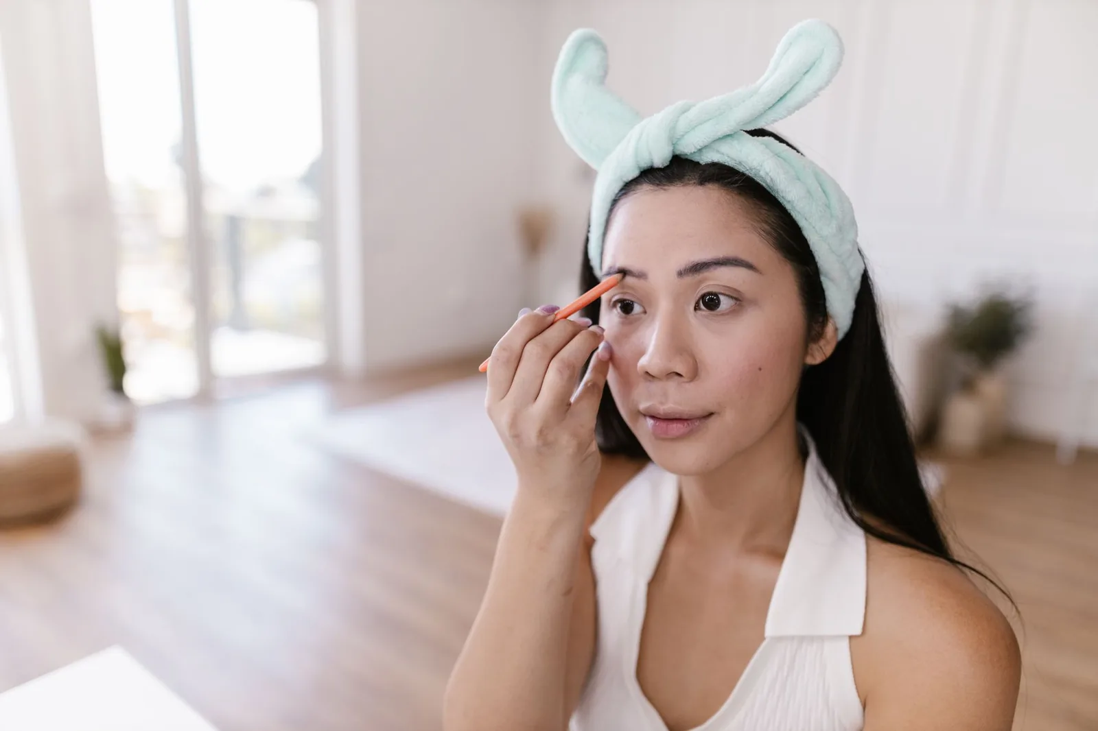 A woman wearing a soft headband fills in her eyebrow with a pencil in a bright room at home.