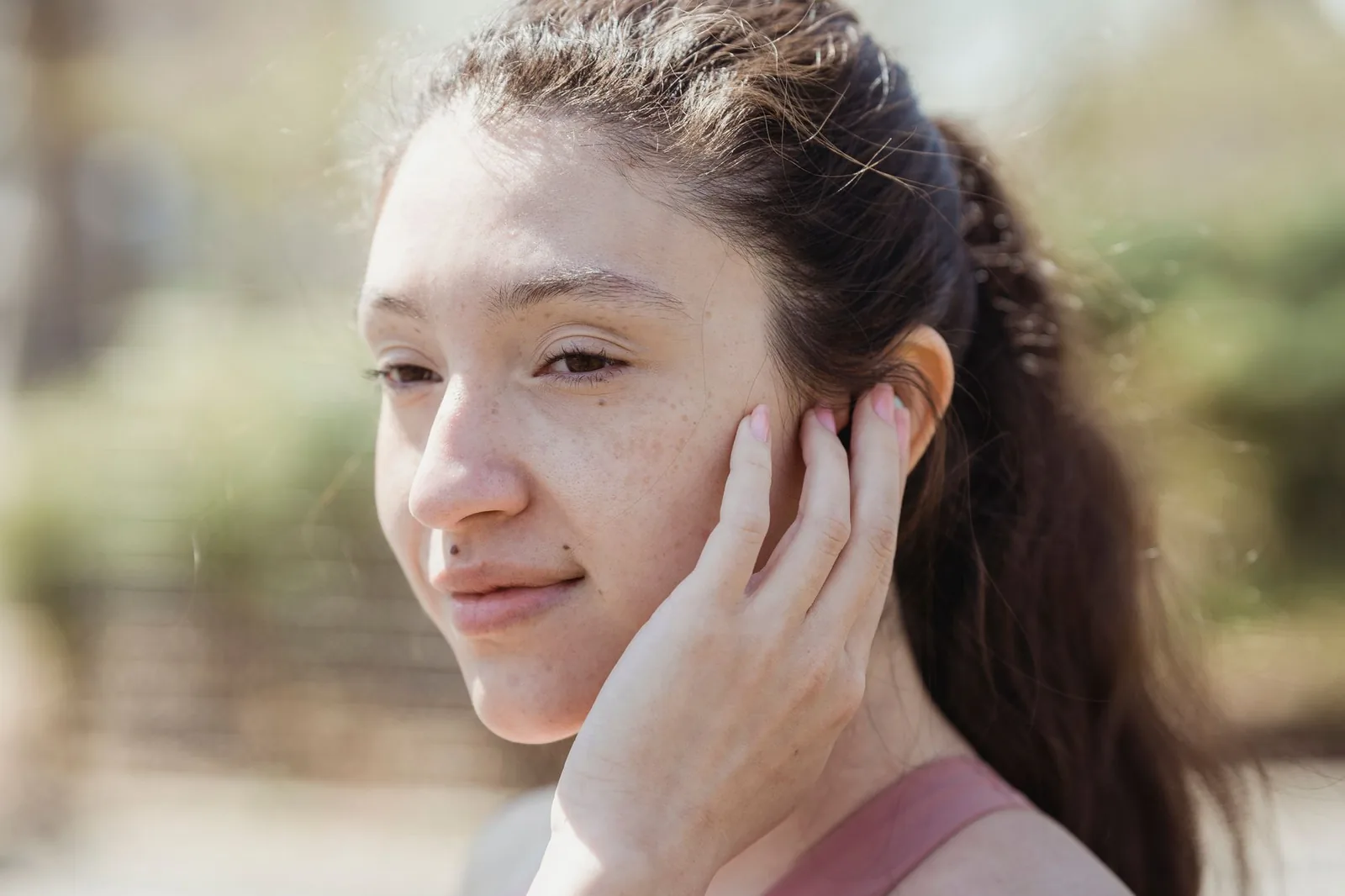 A young woman outdoors resting a hand against her cheek as she checks her own skin.